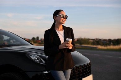 Smiling woman in sunglasses with paper cup of drink near car outdoors