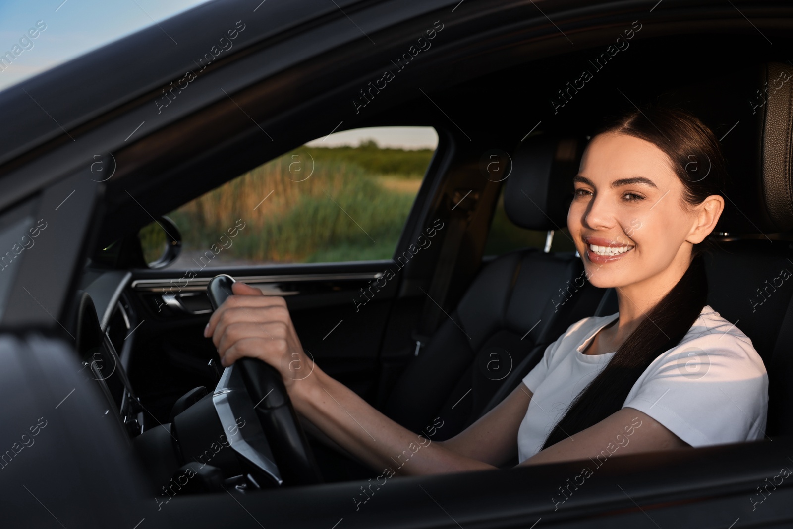 Photo of Smiling young woman holding steering wheel while driving car, view from outside