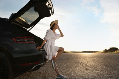 Smiling young woman in hat near trunk of car at sunset, space for text