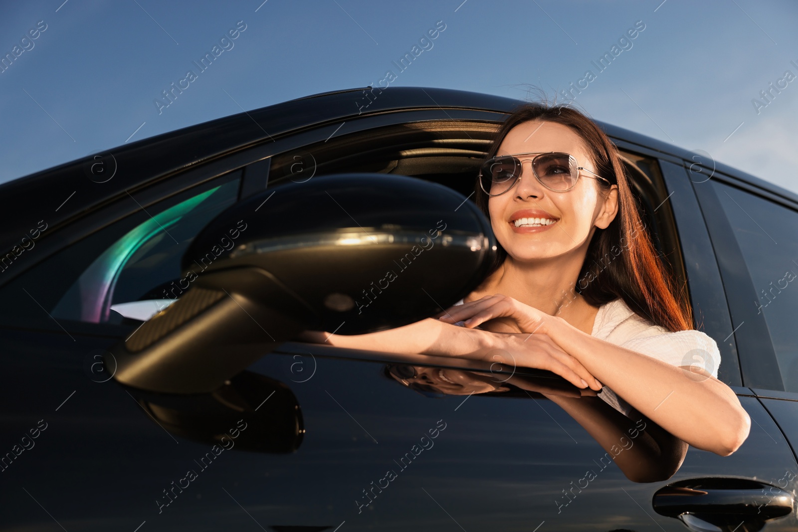 Photo of Smiling young woman in sunglasses leaning out of car window outdoors, low angle view. Enjoying trip