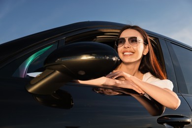 Smiling young woman in sunglasses leaning out of car window outdoors, low angle view. Enjoying trip