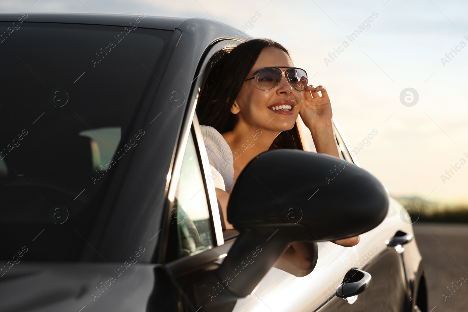 Photo of Smiling young woman in sunglasses leaning out of car window outdoors. Enjoying trip