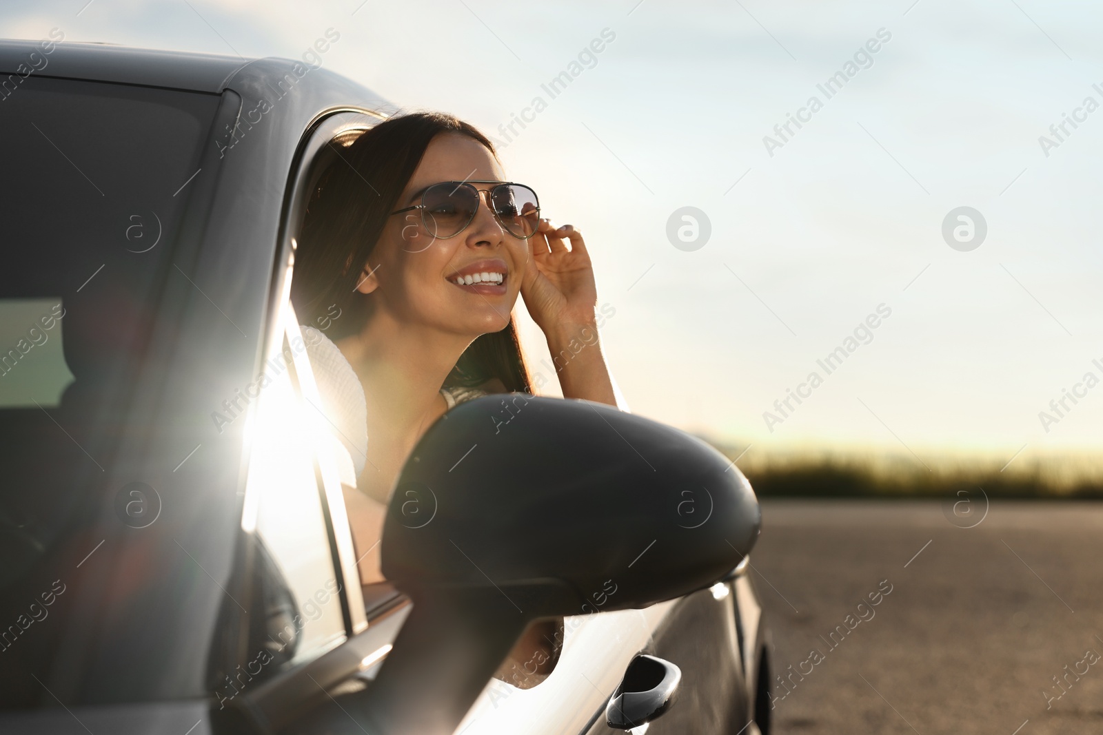 Photo of Smiling young woman in sunglasses leaning out of car window outdoors. Enjoying trip