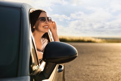 Photo of Smiling young woman in sunglasses leaning out of car window outdoors. Enjoying trip