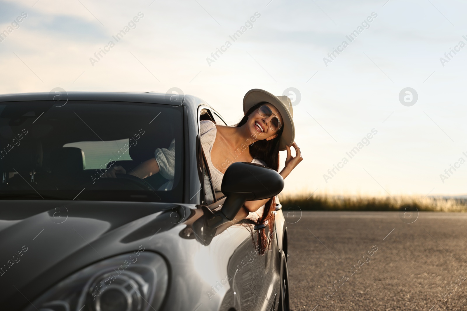 Photo of Smiling young woman in sunglasses with hat leaning out of car window outdoors. Enjoying trip