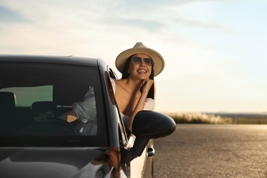 Smiling young woman in sunglasses with hat leaning out of car window outdoors. Enjoying trip