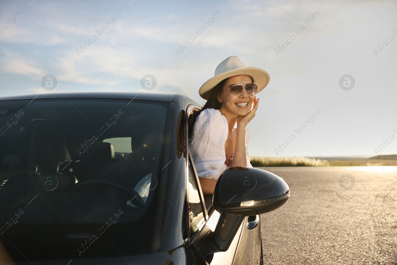 Photo of Smiling young woman in sunglasses with hat leaning out of car window outdoors. Enjoying trip