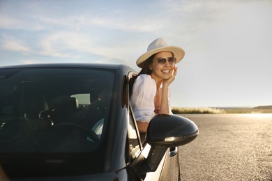 Smiling young woman in sunglasses with hat leaning out of car window outdoors. Enjoying trip