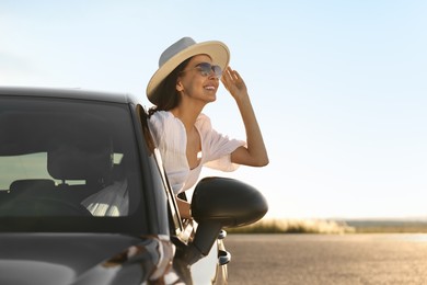 Photo of Smiling young woman in sunglasses with hat leaning out of car window outdoors. Enjoying trip