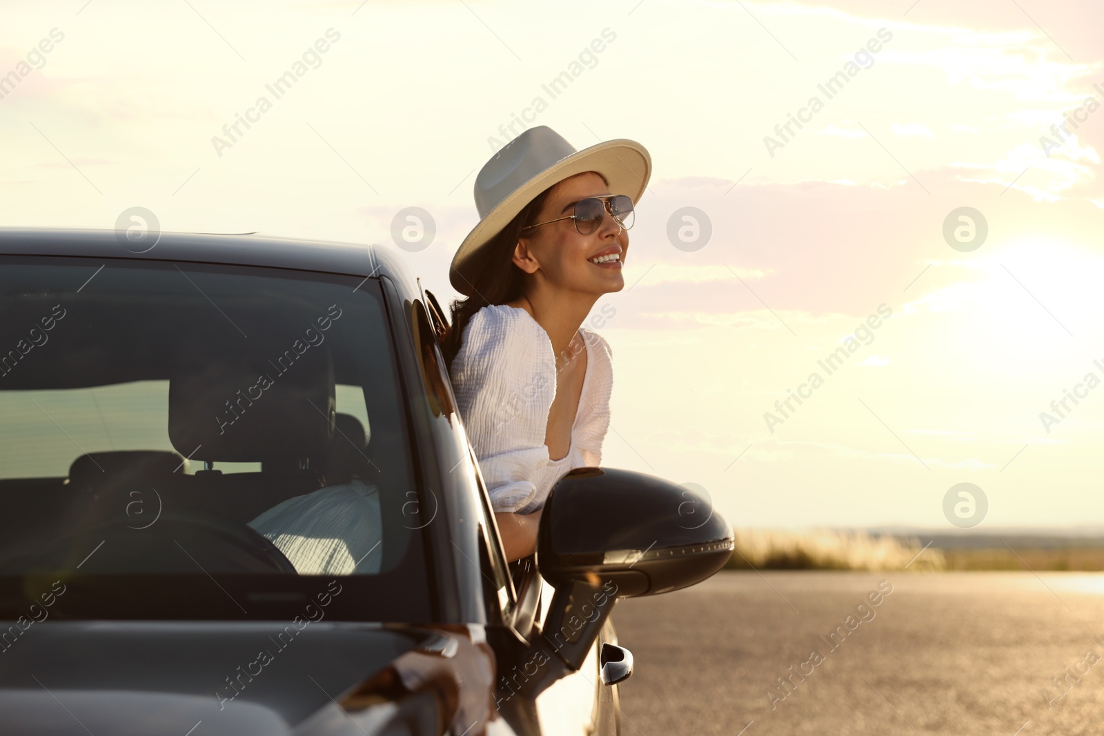 Photo of Smiling young woman in sunglasses with hat leaning out of car window outdoors. Enjoying trip