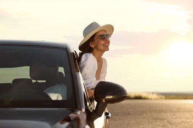 Smiling young woman in sunglasses with hat leaning out of car window outdoors. Enjoying trip