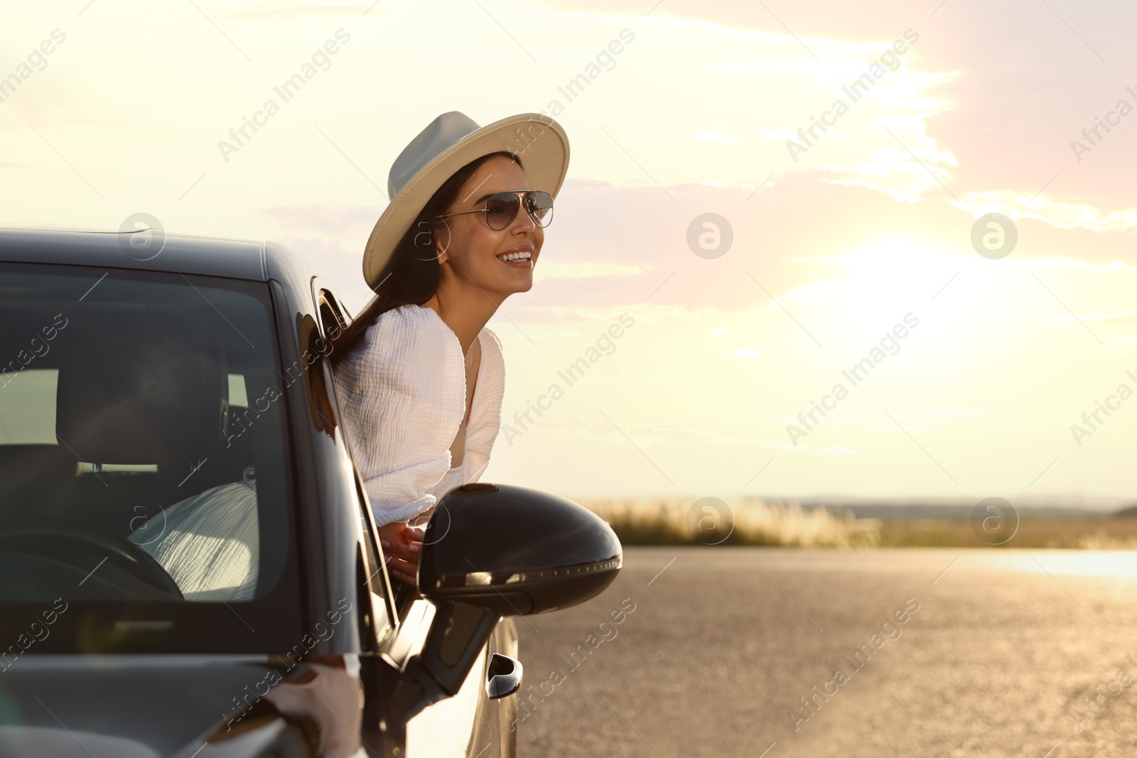 Photo of Smiling young woman in sunglasses with hat leaning out of car window outdoors. Enjoying trip