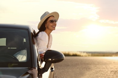 Smiling young woman in sunglasses with hat leaning out of car window outdoors. Enjoying trip