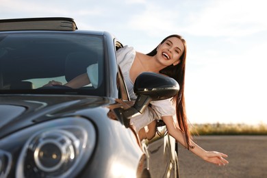 Photo of Smiling young woman leaning out of car window outdoors. Enjoying trip