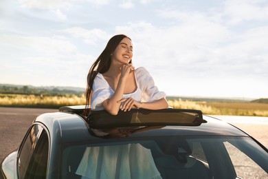 Photo of Enjoying trip. Smiling woman leaning out of car roof outdoors