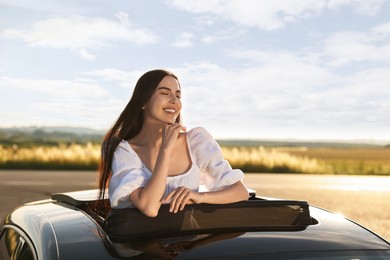 Enjoying trip. Smiling woman leaning out of car roof outdoors