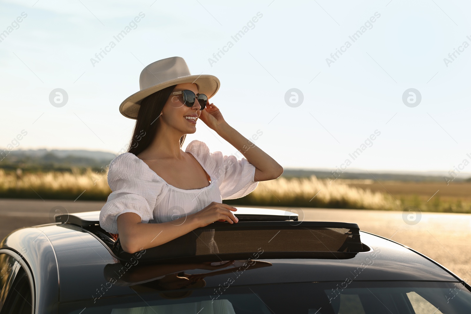 Photo of Enjoying trip. Smiling woman in sunglasses with hat leaning out of car roof outdoors