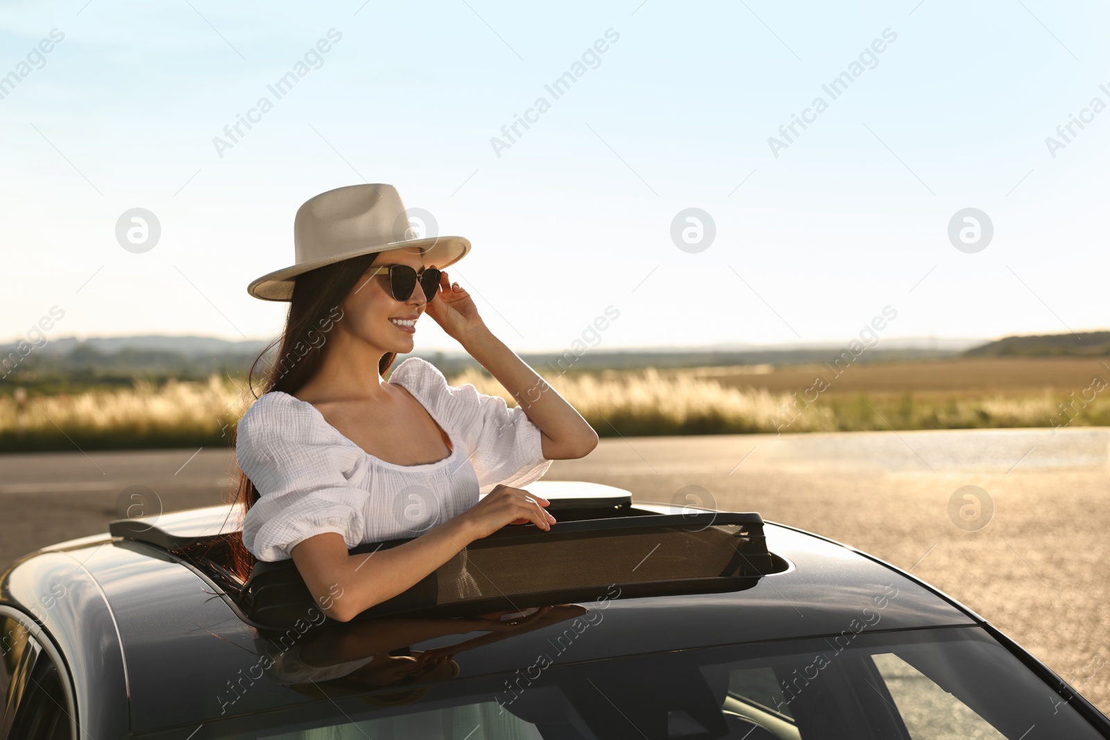 Photo of Enjoying trip. Smiling woman in sunglasses with hat leaning out of car roof outdoors