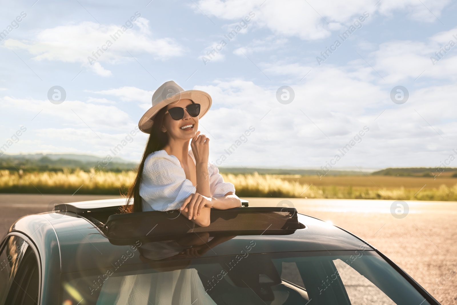 Photo of Enjoying trip. Smiling woman in sunglasses with hat leaning out of car roof outdoors