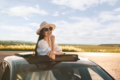 Enjoying trip. Smiling woman in sunglasses with hat leaning out of car roof outdoors