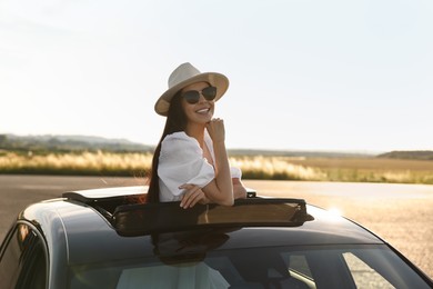 Enjoying trip. Smiling woman in sunglasses with hat leaning out of car roof outdoors