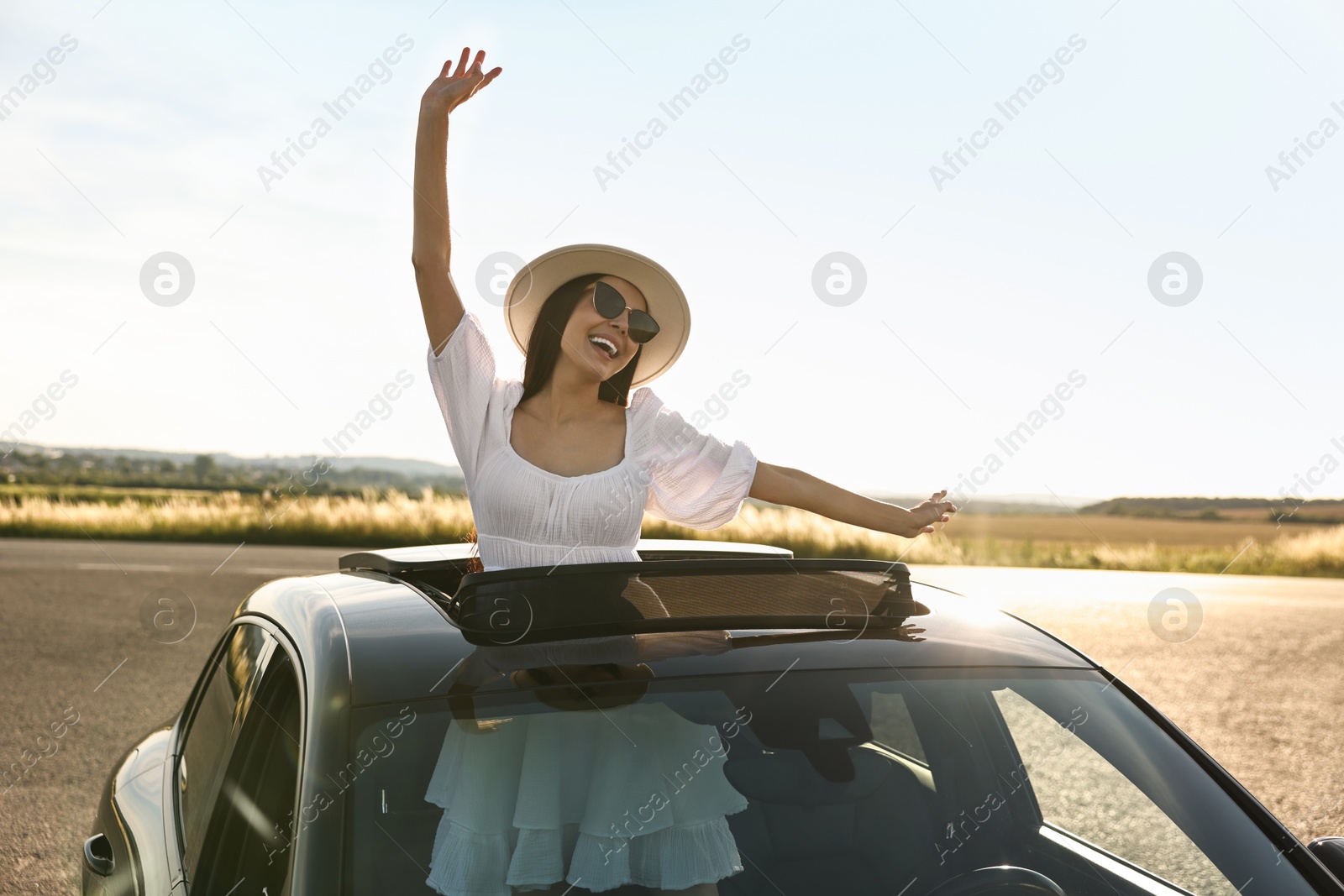 Photo of Enjoying trip. Smiling woman in sunglasses with hat leaning out of car roof outdoors