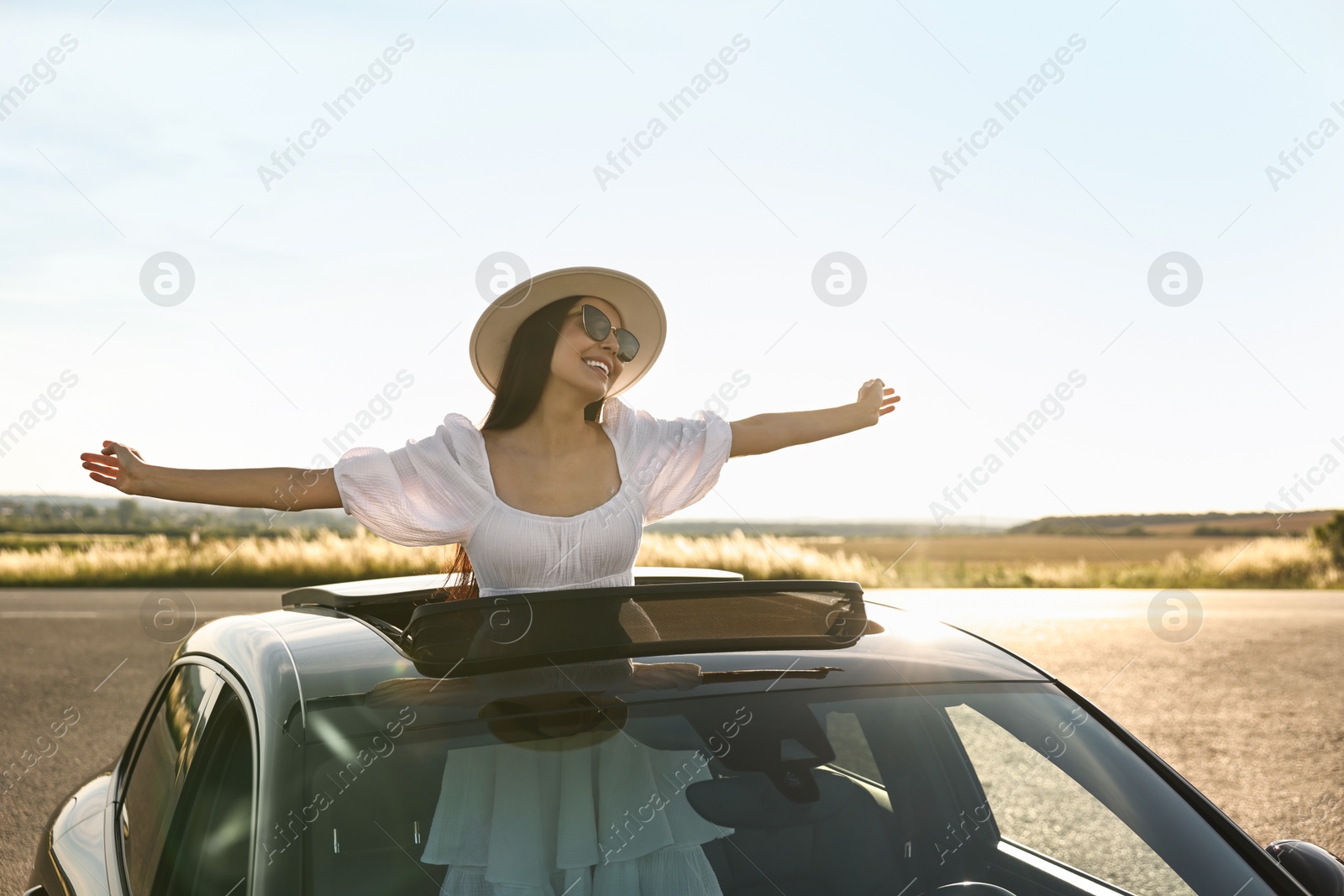 Photo of Enjoying trip. Smiling woman in sunglasses with hat leaning out of car roof outdoors