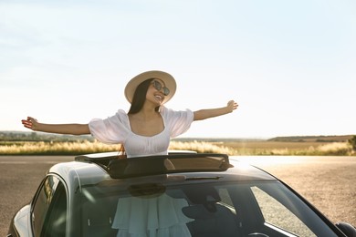 Enjoying trip. Smiling woman in sunglasses with hat leaning out of car roof outdoors