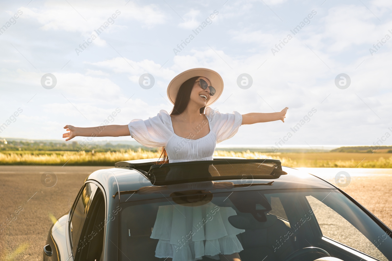 Photo of Enjoying trip. Smiling woman in sunglasses with hat leaning out of car roof outdoors