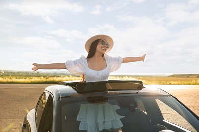 Enjoying trip. Smiling woman in sunglasses with hat leaning out of car roof outdoors