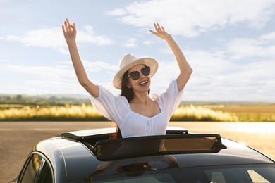 Photo of Enjoying trip. Smiling woman in sunglasses with hat leaning out of car roof outdoors