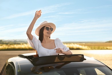 Enjoying trip. Smiling woman in sunglasses with hat leaning out of car roof outdoors