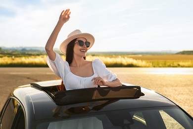 Photo of Enjoying trip. Smiling woman in sunglasses with hat leaning out of car roof outdoors