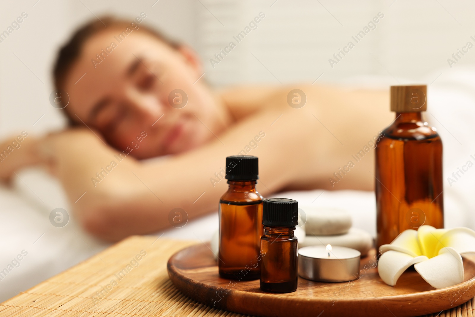 Photo of Aromatherapy. Woman relaxing on massage couch in spa salon, focus on bottles of essential oils, burning candle and plumeria flower