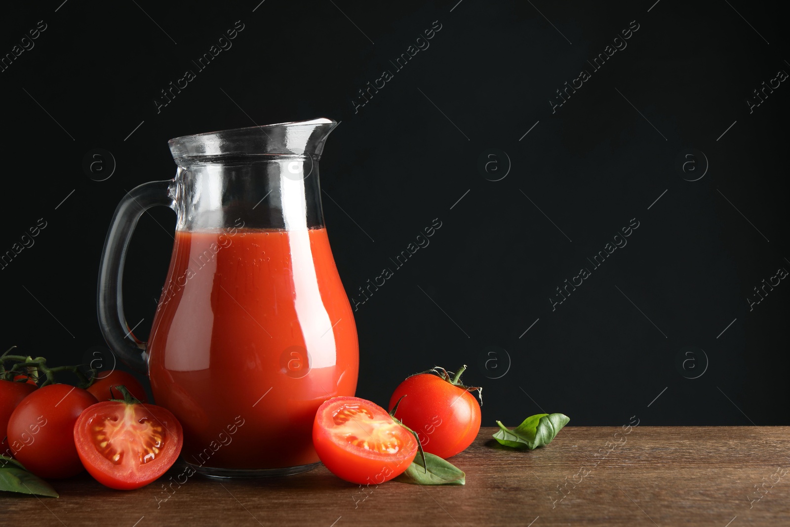 Photo of Tasty tomato juice in jug and vegetables on wooden table against black background. Space for text