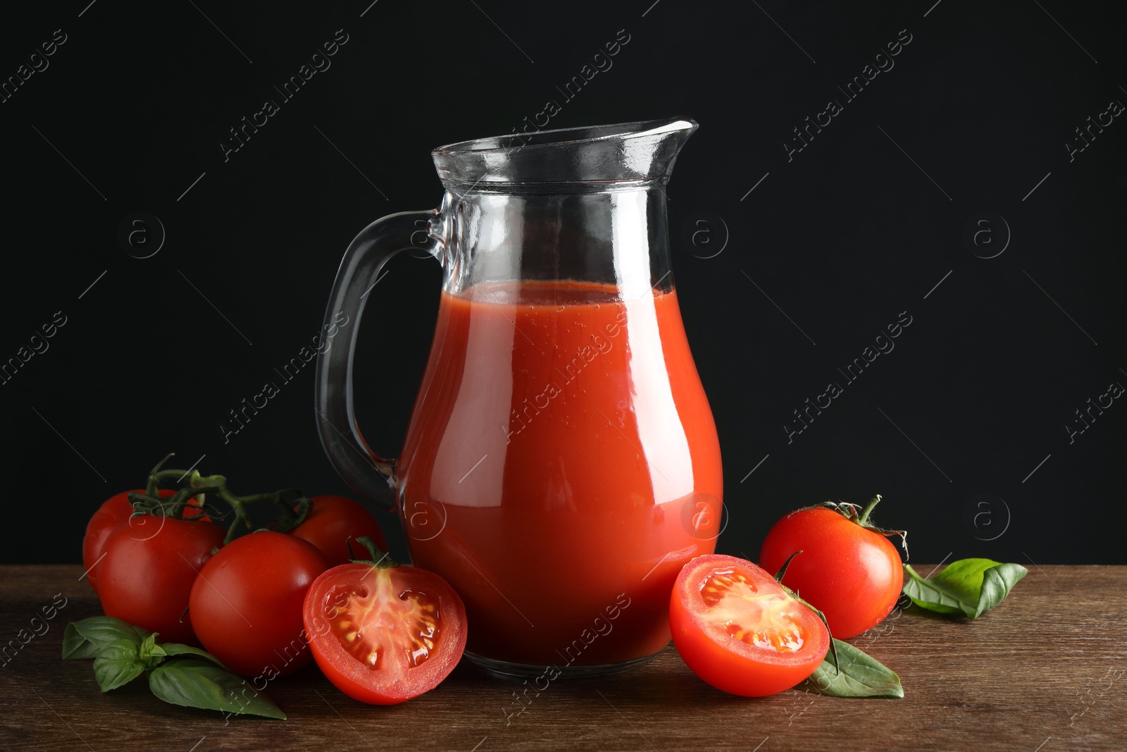 Photo of Tasty tomato juice in jug and vegetables on wooden table against black background