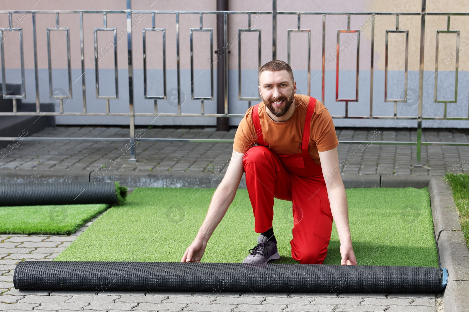 Photo of Happy young man in uniform installing artificial turf outdoors