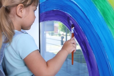 Photo of Little girl drawing rainbow on window indoors