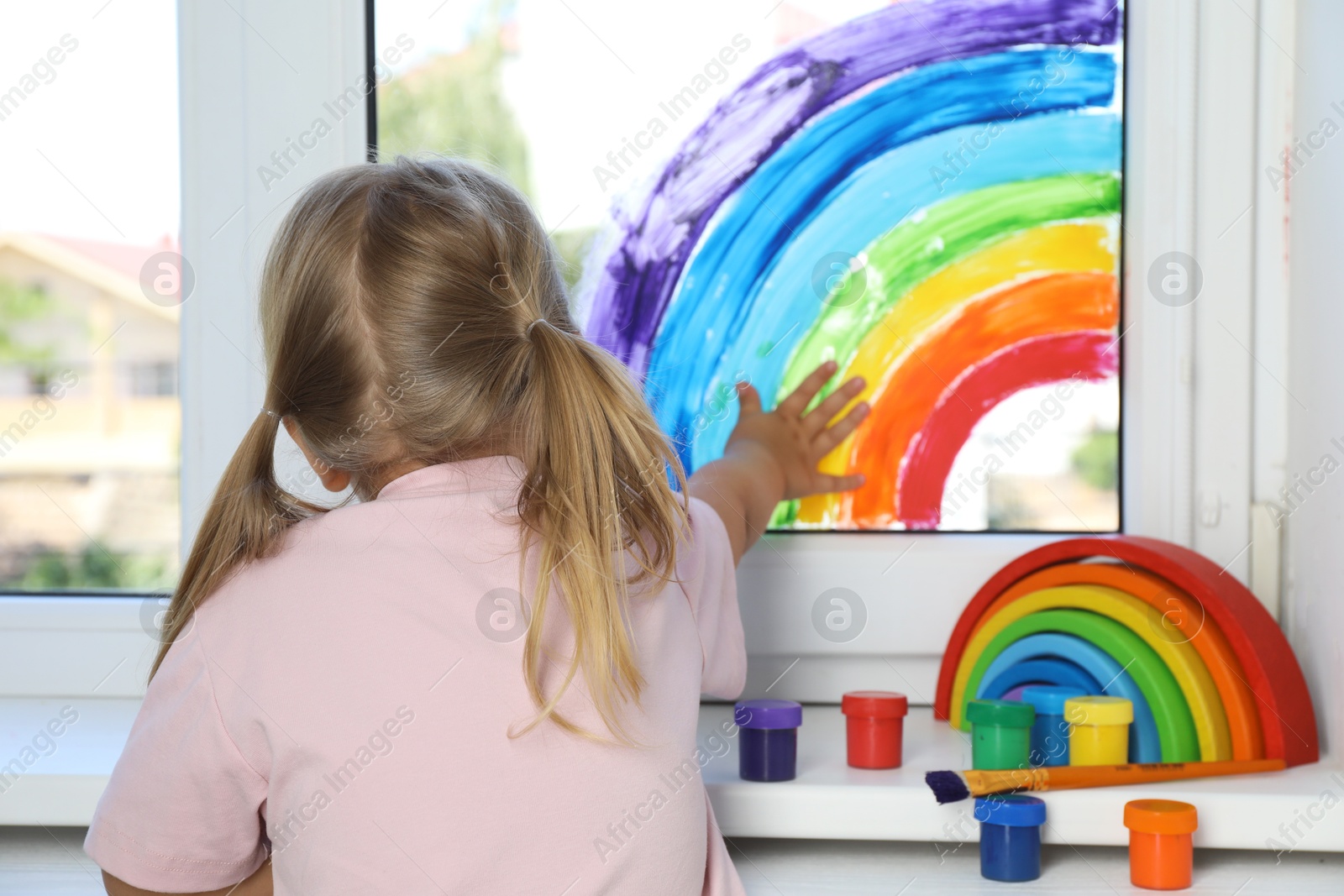 Photo of Little girl touching picture of rainbow on window indoors, back view