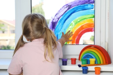 Little girl touching picture of rainbow on window indoors, back view