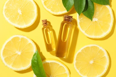 Bottles of essential oils, lemon and green leaves on yellow background, flat lay