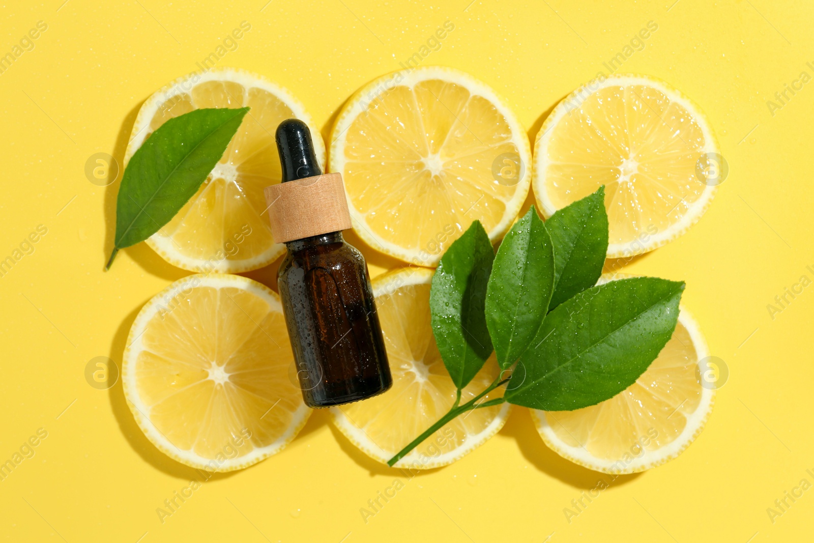 Photo of Bottle of essential oil, lemon and green leaves on yellow background, flat lay