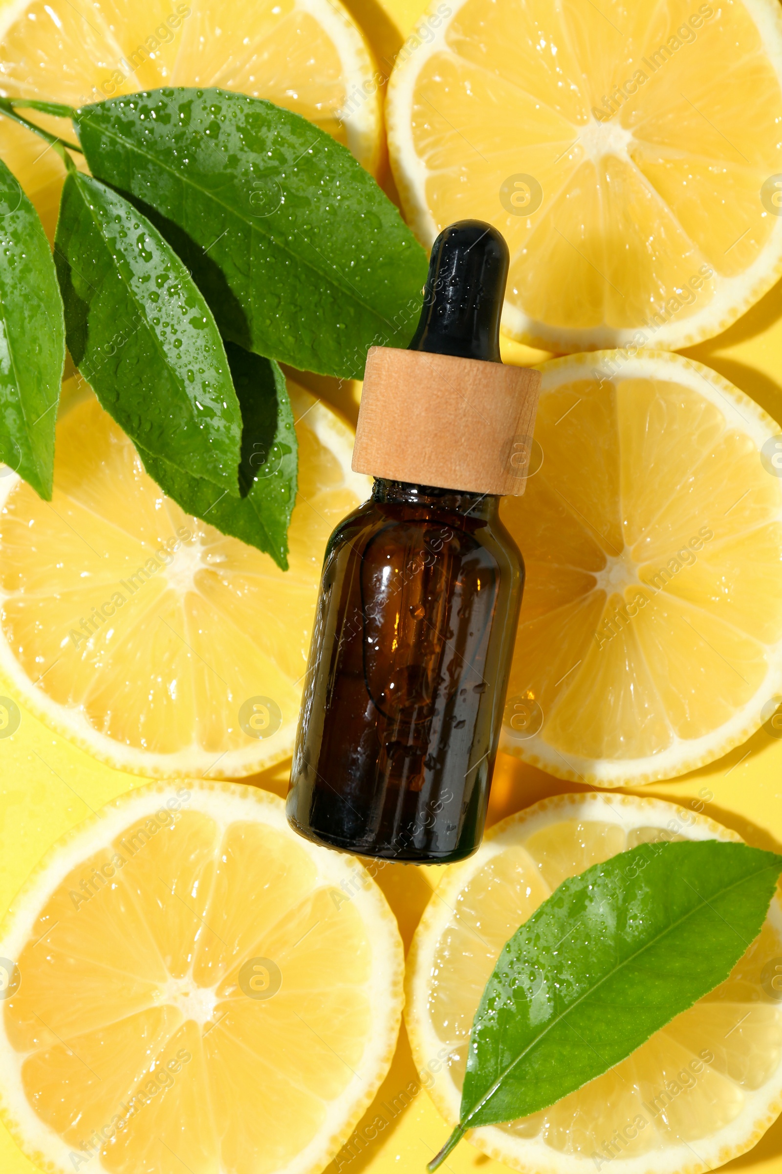 Photo of Bottle of essential oil, lemon and green leaves on yellow background, flat lay