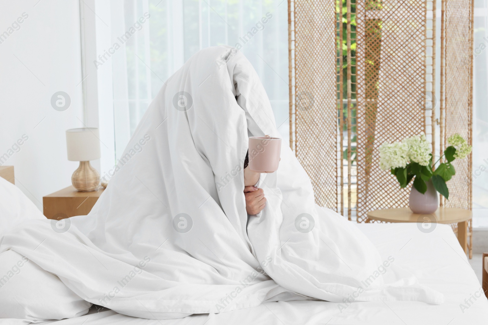 Photo of Bedtime. Woman wrapped in blanket holding cup of drink on bed indoors