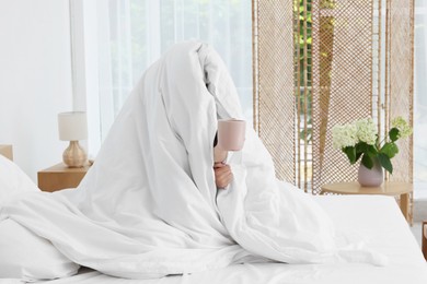 Photo of Bedtime. Woman wrapped in blanket holding cup of drink on bed indoors