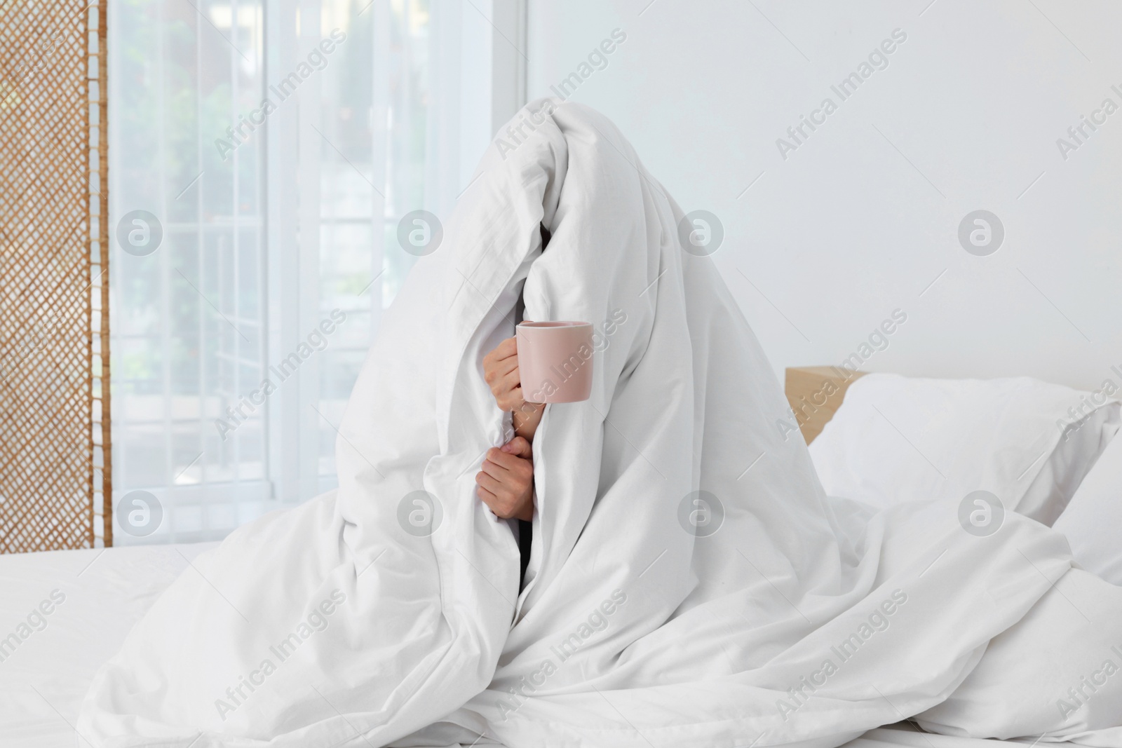 Photo of Bedtime. Woman wrapped in blanket holding cup of drink on bed indoors