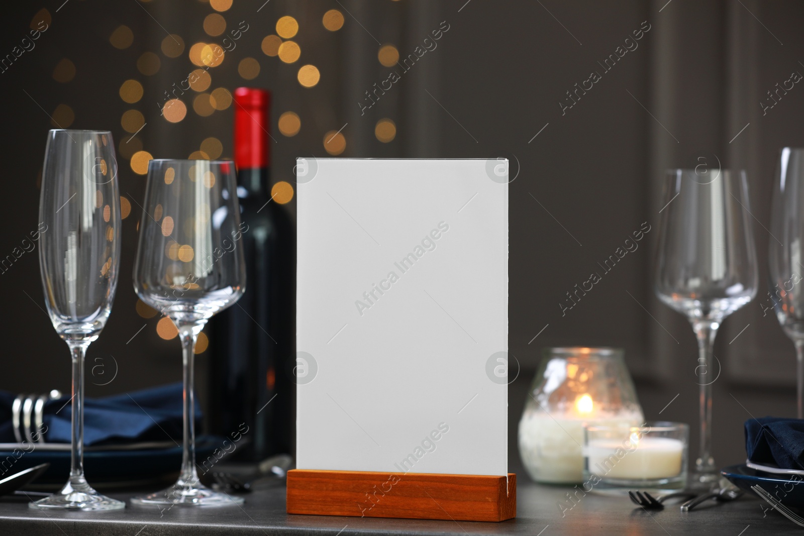 Photo of Menu holder, clean dishware and burning candles on gray table against blurred lights