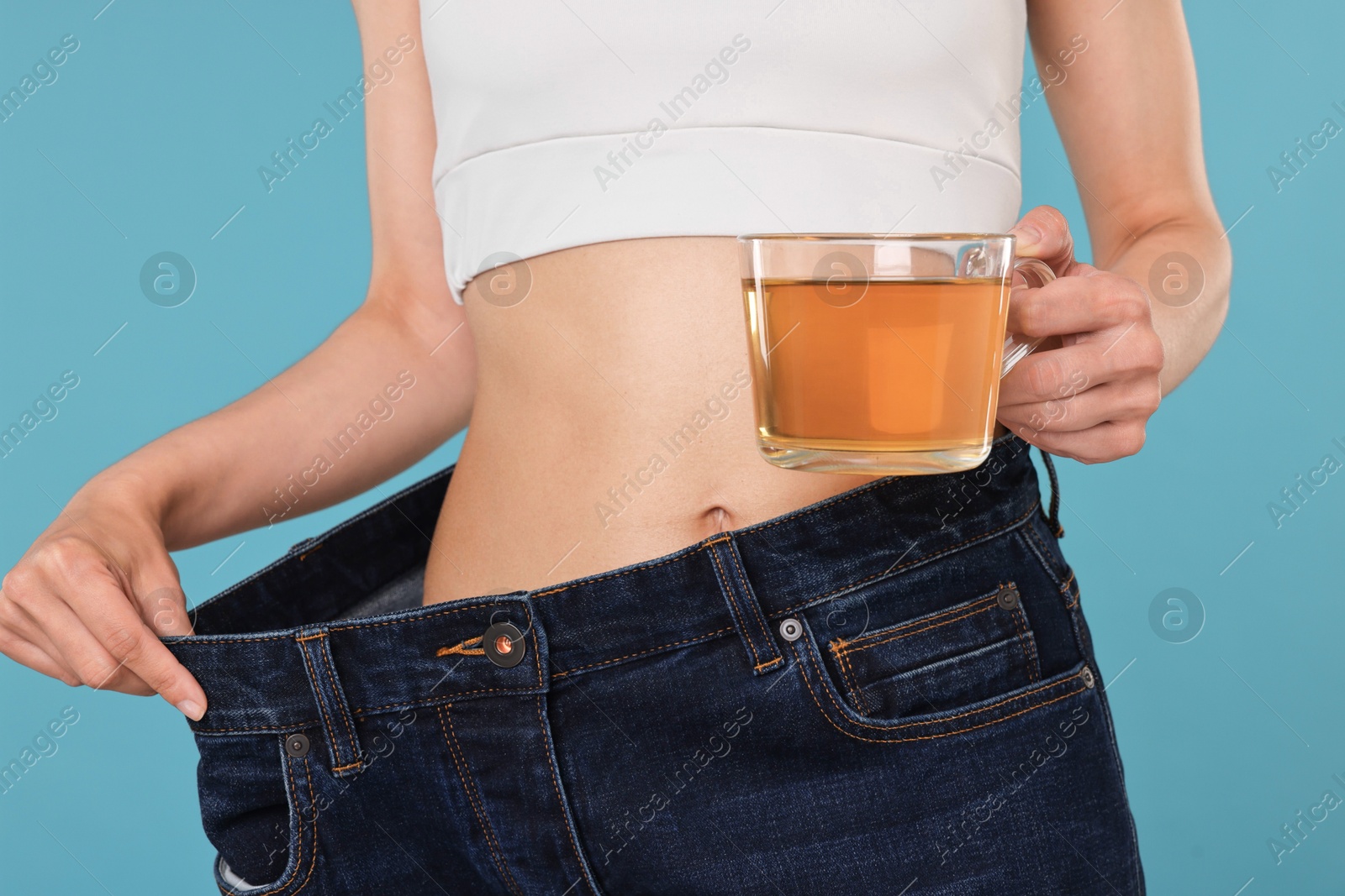 Photo of Weight loss concept. Woman with cup of diet tea and big pants on light blue background, closeup