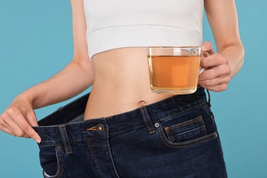 Photo of Weight loss concept. Woman with cup of diet tea and big pants on light blue background, closeup