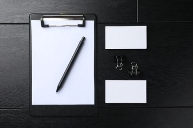 Blank business cards, clipboard with sheet, pen and binder clips on black wooden table, flat lay. Mockup for design